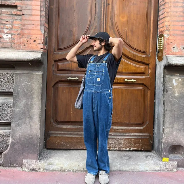 Salopette Carhartt Denim portée par un homme, vue de face en pied, avec t-shirt noir et casquette
