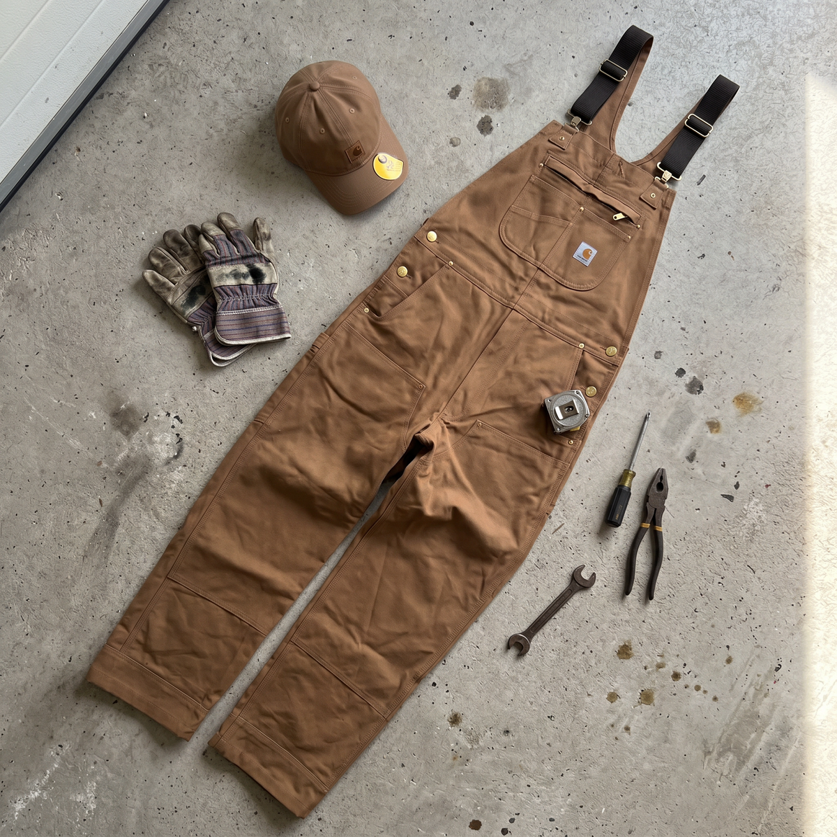 Salopette Carhartt Canvas camel posée à plat, vue de face, avec une casquette, des gants et des outils