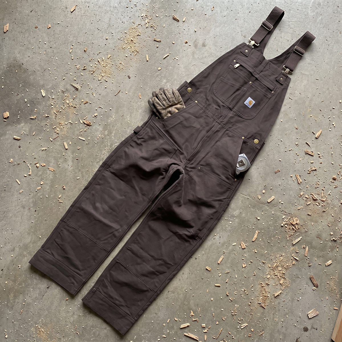 Salopette Carhartt Canvas marron posée à plat, vue de face, avec des gants et un mètre