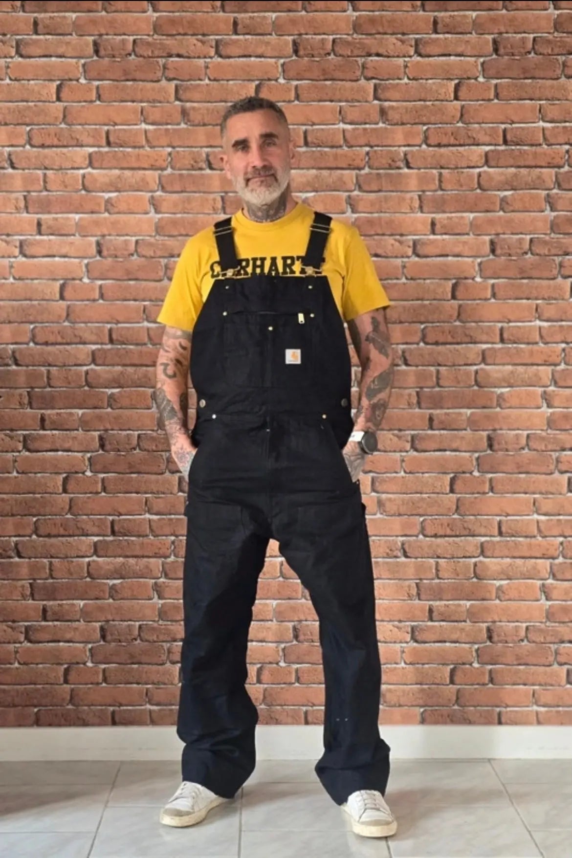 Client en salopette noire et t-shirt jaune devant un mur de briques
