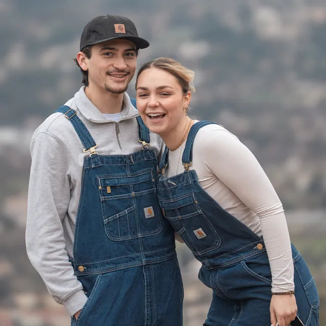 Homme en sweat gris zippé et femme en haut crème à manches longues, tous deux en salopette Carhartt Denim